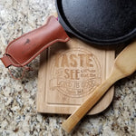 Brown leather handle holder shown on cast iron grill pan with wooden spoon and cutting board